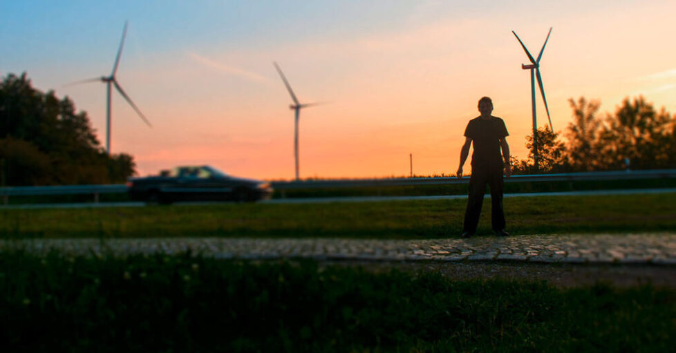 Wiljo Heinen auf einem Rastplatz in der Nähe des Popposteins, Schleswig-Holstein. Im Hintergrund Windräder vor Sonnenuntergang, von links fährt ein Cabriolet ins Bild